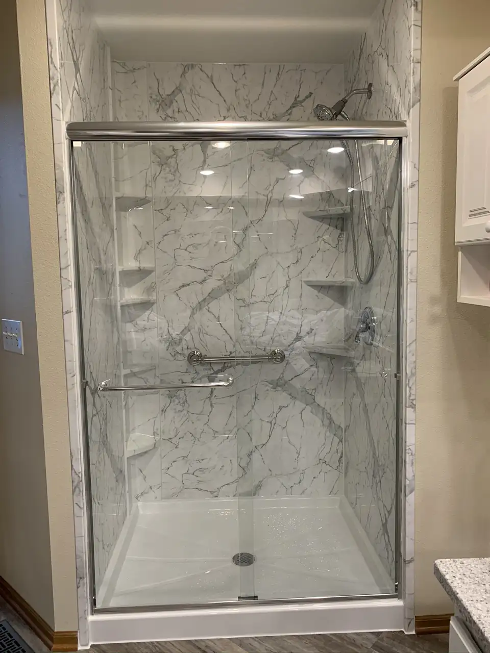 Shower with white marble-look wall panels, corner shelves, a grab bar and a sliding glass door, bathroom remodeling Pleasant Hill